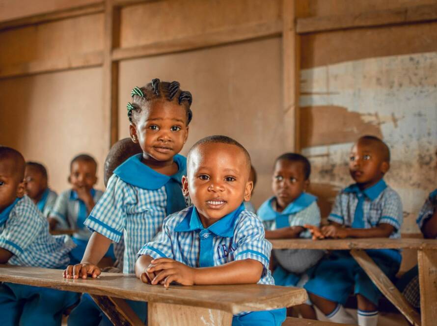 Happy children in a Nigerian classroom wearing uniforms, sitting on wooden benches.